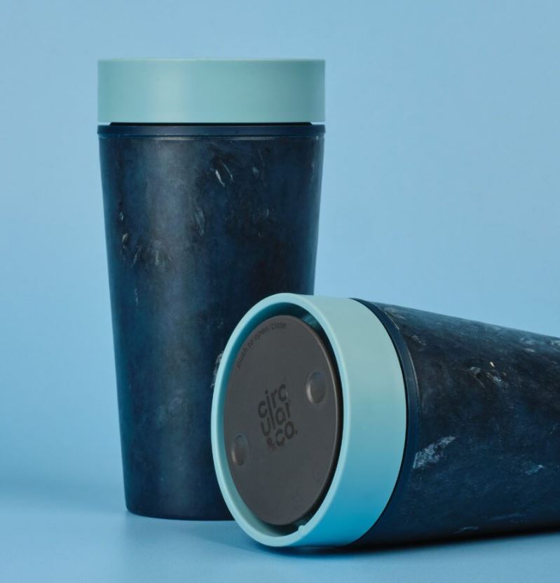 Image of two reusable cups with dark blue cups and light blue lids, against a light blue background. One cup is standing upright, whilst the other is laying horizontally showing the top of the cup. 