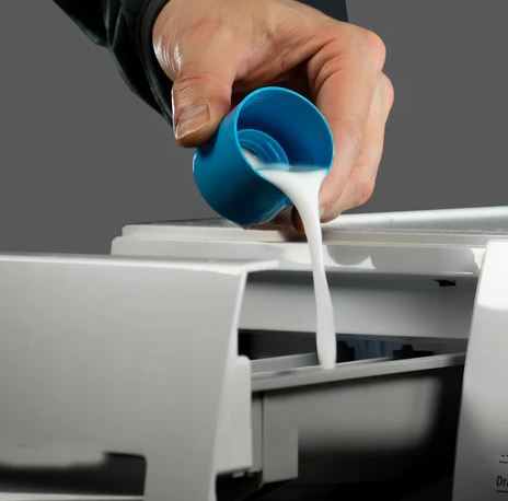 Image showing a hand pouring liquid detergent from a blue measuring cup into a washing machine detergent drawer. 