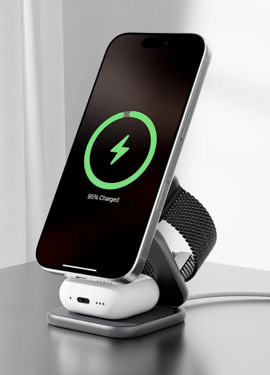 Image of smartphone, wireless earbuds and smartwatch placed on a dark grey/silver multi-device charging station. The smartphone displays a green charging logo. 
