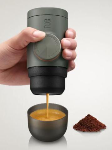 hand holding portable grey coffee machine, with coffee pouring into the cup below and a pile of coffee ground to the site 