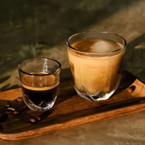 two glass cups on a wooden board, the smaller glass at the front has an espresso in it and the larger one at the back has a latte in it