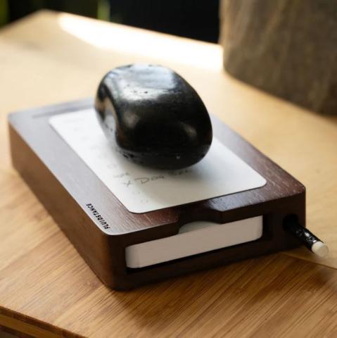 wooden block with a card on top, with a black stone on it. All on a wooden desk. 