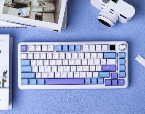 white keyboard with light blue, white and lilac keys. The keyboard has a rotary knob and a mini screen n the top right corner. The keyboard in on a lilac background with white accessories such as a camera and some books next to it. 