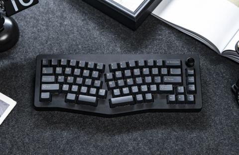 Black keyboard with keys divided into two halves to support better wrist support. The keyboard is on a dark grey surface with the edge of a notebook in the top right corner