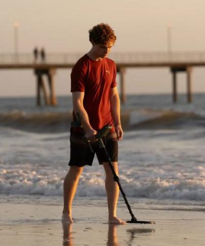 man in a red tshirt and black shorts walking on beach with the sea behind him, holding a metal detector above the ground 