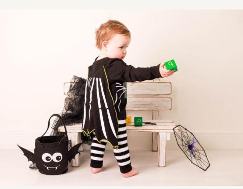 A small child playing with a green ball wearing black and white striped leggings, a black top which has a black and white striped cape attached to it. There is also a black spider bag sat next to the child on on the floor. 
