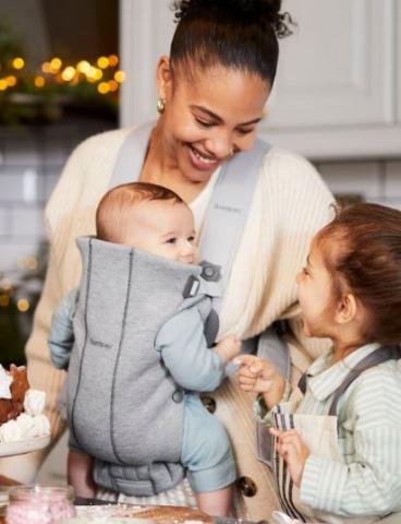 woman wearing a baby carrier with a baby in it, with a young child next to her. Both the woman and the young child are looking at the baby in the carrier. 