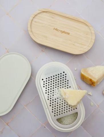 Grater with container on a grey table with a plastic lid and wooden lid next to it. On top of the grater is a block of cheese and some cheese shavings, with another bit of cheese next to the grater too. 
