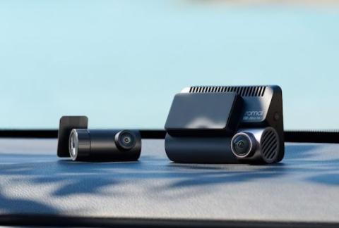 Photograph of two compact dash cameras placed on a car dashboard against a blurred blue background. One camera is cylindrical with a lens on one end, and the other is rectangular with a lens and ventilation slits, featuring the brand name "70mai" on its side.