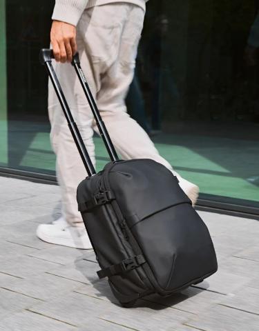 Person pulling a black wheeled suitcase/rucksack with an extension handle on a cement flood and near a glass wall