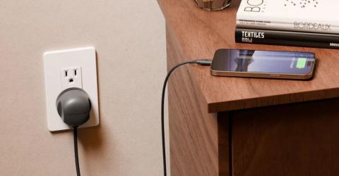 phone on a wooden table being charged by a grey charging cable and plug adapter, plugged into a white wall socket 