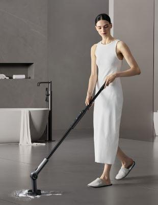 A person in a white sleeveless dress and slippers using a black cordless spinner scrubber tool to clean a light-grey floor in a modern bathroom setting. The scene features a freestanding bathtub with a towel draped over the edge and a minimalist gray wall background.