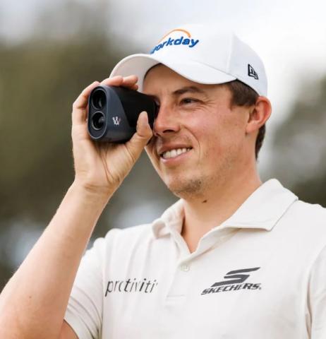 Image of a person wearing a white polo shirt and cap, holding a black rangefinder device up to their eye. 