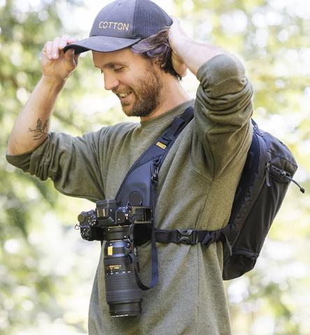 Image of person adjusting a dark grey cap, wearing a green long-sleeve shirt and a black bag over one shoulder. The bag has a professional camera attached to the front on the bag's chest harness strap. 