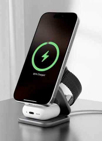 Image of smartphone, wireless earbuds and smartwatch placed on a dark grey/silver multi-device charging station. The smartphone displays a green charging logo. 