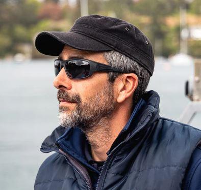 Image of man wearing a black cap, black wrap around sunglasses and a navy puffer vest. The background of the image shows a blurred body of water and trees in the landscape. 
