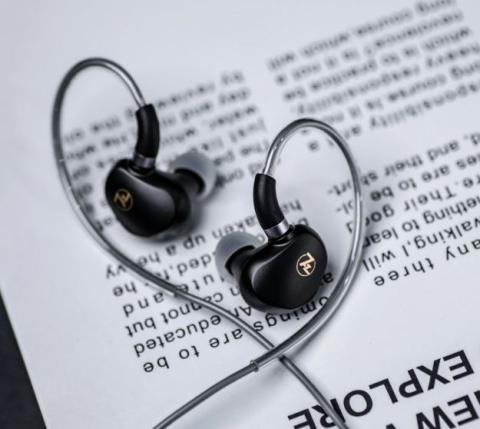 Image of black in-ear monitors with silver cable, on a white background with black text which is slightly blurred on it. 