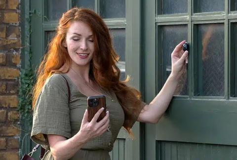 Image of woman with long red hair and wearing a green top standing in front of a green door. In one hand the woman is holding a phone, and in the other they are pressing the Nix Sensor Mini 3 against the green door. 