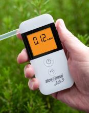 white breathalyzer held in hand with a clear straw at the side of it with an orange screen displaying alcohol level 
