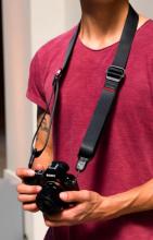 person in a red tshirt with a DSLR black camera on a black camera strap which is around the persons neck 
