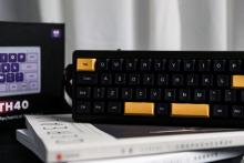 small black and orange keyboard with a lanyard stood up on a stack of books with the keyboard's black and purple box behind it 