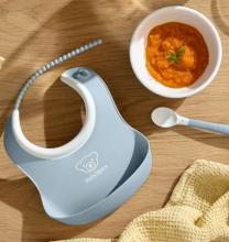 a blue and white babys bip, bowl and spoon on a wooden table. The bowl contains an orange puree with a green garnish on top. 