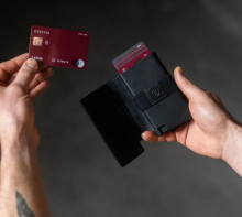 hands holding a black card holder with a black leather flap. The cardholder has red cards in it. The other hand is holding a red credit card. 