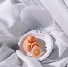 peach sleep earplugs in a white circular case that is open, on a white fabric bedsheet-like material background