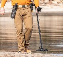 waist down image of person wearing yellow jumper, beige trousers and brown boots walking on the beach with a black metal detector in one hand, the beach background shows shallow water and sand