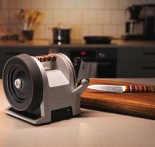 silver and black knife sharpener unit with a colourful striped top handle at the top of it, on a kitchen side, with a knife on a wooden chopping board next to to it