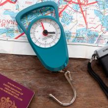 blue travel scale with scale face and bottom metal hook, on a wooden table with a map in the background and a passport in the bottom left corner 