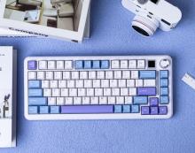 white keyboard with light blue, white and lilac keys. The keyboard has a rotary knob and a mini screen n the top right corner. The keyboard in on a lilac background with white accessories such as a camera and some books next to it. 