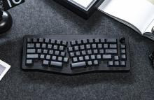 Black keyboard with keys divided into two halves to support better wrist support. The keyboard is on a dark grey surface with the edge of a notebook in the top right corner