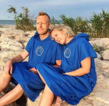a man and woman sat on a rock on the beach, both are wearing blue changing robes, and the woman has her head on the mans shoulder 