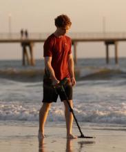 man in a red tshirt and black shorts walking on beach with the sea behind him, holding a metal detector above the ground 