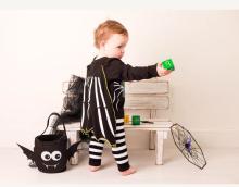 A small child playing with a green ball wearing black and white striped leggings, a black top which has a black and white striped cape attached to it. There is also a black spider bag sat next to the child on on the floor. 