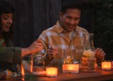 two people sat at a bench which has food and three cube lanterns on the table top. The lights are emitting a warm light across the setting. 