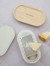 Grater with container on a grey table with a plastic lid and wooden lid next to it. On top of the grater is a block of cheese and some cheese shavings, with another bit of cheese next to the grater too. 