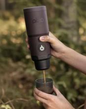 Hands holding a cylinder shaped coffee machine above a coffee cup with coffee being poured into the cup from the machine. Image on a blurred forest background. 