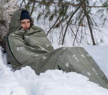 man wearing sleeping-bag style tent in green, sat in the snow against a tree