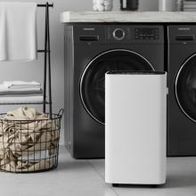 white square dehumidifier unit in front of a grey washing machine and next to a basket with clothes in it