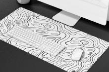 Computer set up with a white keyboard and mouse on a large desk mat with black contour line patterns resembling a topographic map. 