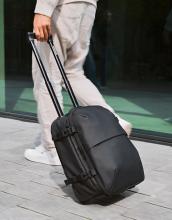 Person pulling a black wheeled suitcase/rucksack with an extension handle on a cement flood and near a glass wall