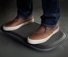 Photograph showing a person wearing brown leather shoes and dark blue jeans standing on a black balance board with a textured hexagonal pattern.