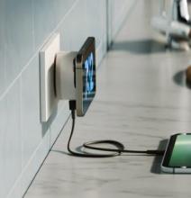 phone attached to white wall socket charging unit, charging unit also has a cable at the base charging a tablet that is at the edge of the image