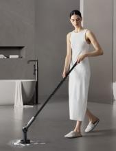 A person in a white sleeveless dress and slippers using a black cordless spinner scrubber tool to clean a light-grey floor in a modern bathroom setting. The scene features a freestanding bathtub with a towel draped over the edge and a minimalist gray wall background.