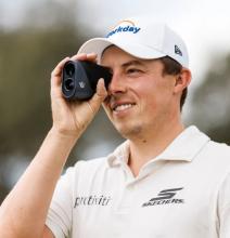 Image of a person wearing a white polo shirt and cap, holding a black rangefinder device up to their eye. 