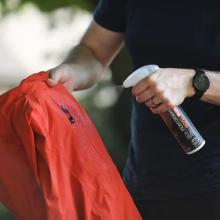 Image Showing a person spraying a red jacket with a waterproofing product labelled 'Performance Repel Plus'. 
