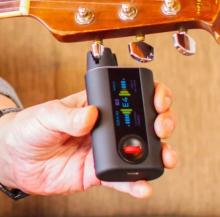 Image showing a hand holding a black digital guitar tuner clipped to an acoustic guitar peg.