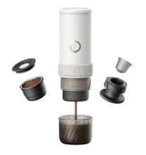 Image of a white cylindrical portable espresso maker with components on either side, including a ground coffee basket and a coffee pod holder. At the base is a glass cup catching brewed coffee. 